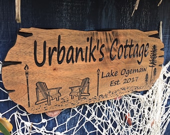 May include: A wooden sign engraved with "Urbanik's Cottage Lake Ogemaw Est. 2017". The sign includes illustrations of chairs, a torch, and pine trees. The sign is brown with a natural edge, and is likely for a lakeside cottage.