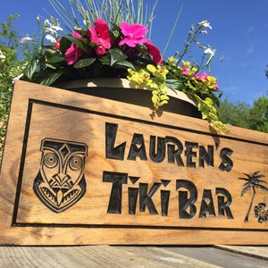 Pode incluir: Placa de madeira com a inscri&ccedil;&atilde;o "Lauren's Tiki Bar", uma m&aacute;scara tiki, uma palmeira e uma flor. A placa est&aacute; colocada em frente a uma floreira com flores cor-de-rosa.