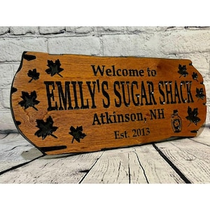 May include: A wooden sign with the text "Welcome to Emily's Sugar Shack, Atkinson, NH Est. 2013". The sign is decorated with maple leaves and a silhouette of a maple syrup bottle.