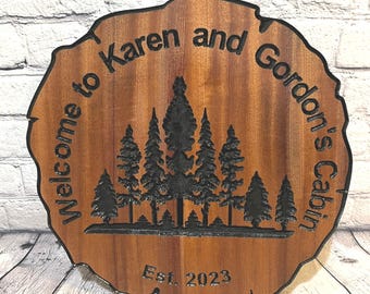 May include: Welcome to Karen and Gordon's Cabin, a wooden sign with a rustic woodgrain finish, featuring a silhouette of a forest with pine trees and the text "Welcome to Karen and Gordon's Cabin Est. 2023".