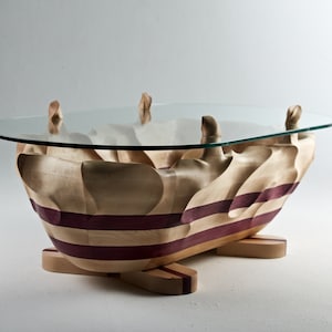 Op de afbeelding: Een salontafel met een helder ovaal glazen blad op een sculpturale houten basis. De basis is gemaakt van lichtgekleurd hout met horizontale banden van donkerpaars hout, voorzien van organische, vloeiende rondingen en vier kleine houten voetjes.