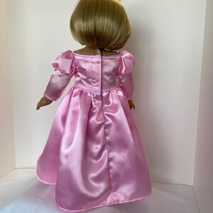 May include: A doll with short blonde hair, wearing a light pink satin dress. The dress has long sleeves with puffed shoulders and a full skirt. The back of the dress has a white zipper and a small bow at the waist. The doll is standing against a white background.