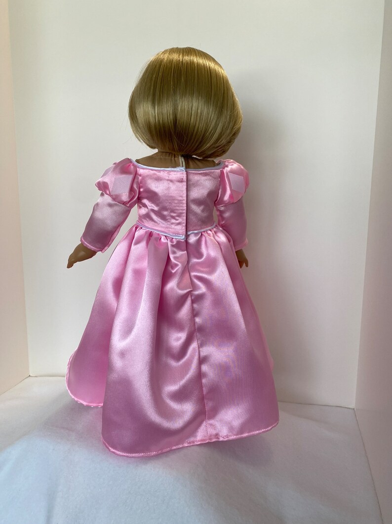 May include: A doll with short blonde hair, wearing a light pink satin dress. The dress has long sleeves with puffed shoulders and a full skirt. The back of the dress has a white zipper and a small bow at the waist. The doll is standing against a white background.