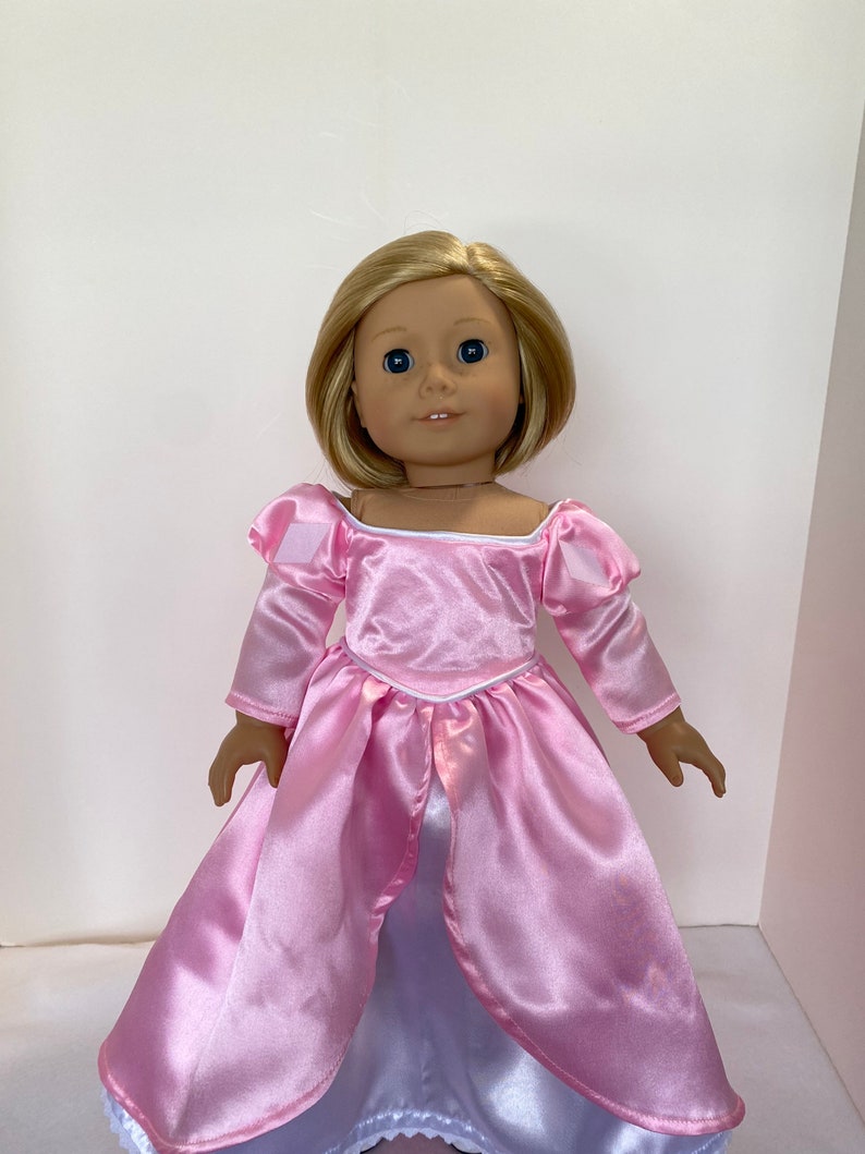 May include: A doll with blonde hair and blue eyes wearing a pink satin dress with long sleeves and a white underskirt. The dress has a square neckline and a fitted bodice.