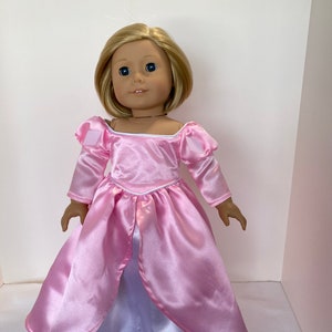 May include: A doll with blonde hair and blue eyes wearing a pink satin princess dress. The dress has long sleeves, a square neckline, and a white underskirt with a lace trim. The doll is standing upright.