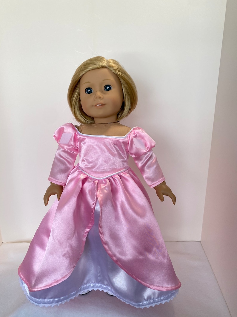 May include: A doll with blonde hair and blue eyes wearing a pink satin princess dress. The dress has long sleeves, a square neckline, and a white underskirt with a lace trim. The doll is standing upright.