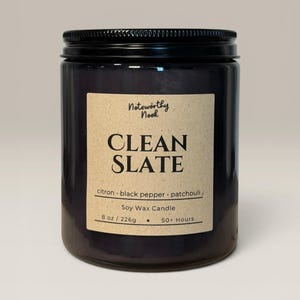 May include: A black glass jar soy wax candle with a black screw-top lid. The beige label features the text Noteworthy Nook, CLEAN SLATE, citron, black pepper, patchouli, Soy Wax Candle, 8 oz / 226g, and 50+ Hours in black serif and sans-serif fonts.