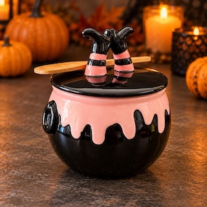 May include: A black and pink ceramic cauldron-shaped jar with a lid featuring two legs wearing black and pink striped stockings. The jar has a glossy finish with pink drip-style accents at the top and includes a wooden spoon resting on the lid.