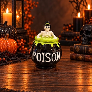 May include: A black ceramic jar shaped like a cauldron with the word POISON printed in white letters. The lid is bright green with a sculpted white skeleton wearing a small black top hat emerging from the center of the pot.