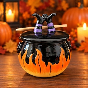 May include: A black ceramic cauldron with a lid featuring a pair of black and purple striped legs sticking out of the top. The base of the cauldron is decorated with painted orange and yellow flames. A small wooden spoon rests across the lid.