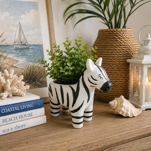 May include: A white ceramic planter shaped like a standing zebra with black stripes and a black nose. The planter holds a small green leafy plant. The zebra has closed eyes with eyelashes painted on its face and stands on four short legs.
