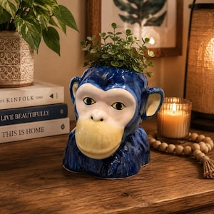 May include: A glossy ceramic monkey-shaped planter featuring a deep blue finish on the head and shoulders, with a contrasting pale yellow muzzle, dark eyes, and a small green plant growing from the top opening of the head.