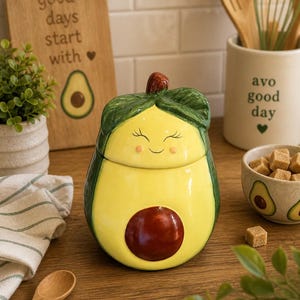 May include: A ceramic avocado-shaped canister with a removable lid. The canister features a glossy yellow center, a dark brown pit, and a green top with textured leaves. The lid has a painted face with closed eyes, a small smile, and pink cheeks.