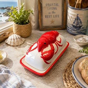 May include: A white ceramic butter dish with a red lobster figurine on the lid. The rectangular base has a red rim. The lobster is glossy red with black eyes and detailed claws. The dish is designed to hold a stick of butter for table service.