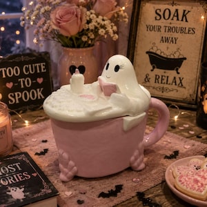May include: A ceramic mug shaped like a pink clawfoot bathtub with a white ghost figure sitting inside. The ghost is reading a small book and has a candle nearby. The mug features a textured bubble surface on top and a curved handle on the side.
