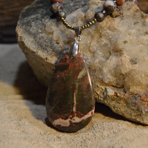May include: A teardrop-shaped pendant necklace with a brown, green, and pink stone. The stone is set in a silver pendant and hangs from a brown beaded chain.