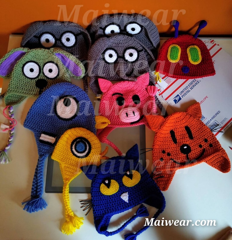 May include: A collection of colorful crocheted hats with different animal faces. The hats feature a variety of characters, including a cat, a pig, a bug, a dog, and a monster. Each hat has a long tail and is made with a soft, textured yarn.