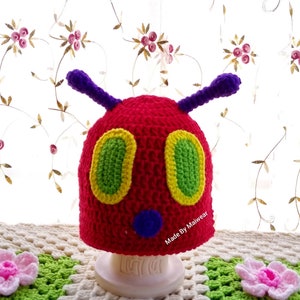 May include: A red, green, and yellow crocheted hat shaped like a caterpillar with purple antennas and a blue face. The hat is sitting on a white stand. The text "Made By Maiwear" is on the hat.