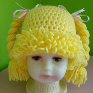 May include: A yellow crocheted hat with pigtails and a fringe trim. The hat is on a white mannequin head.