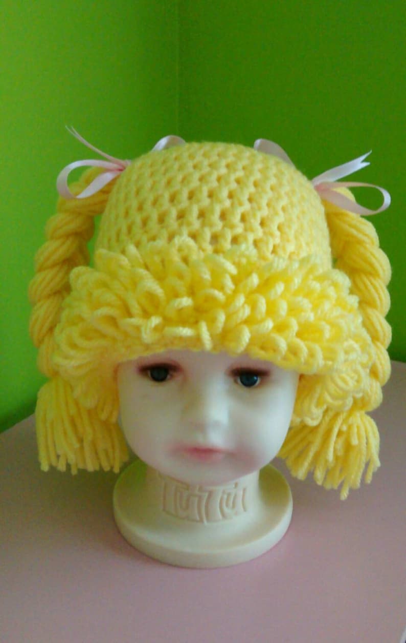 May include: A yellow crocheted hat with pigtails and a fringe trim. The hat is on a white mannequin head.
