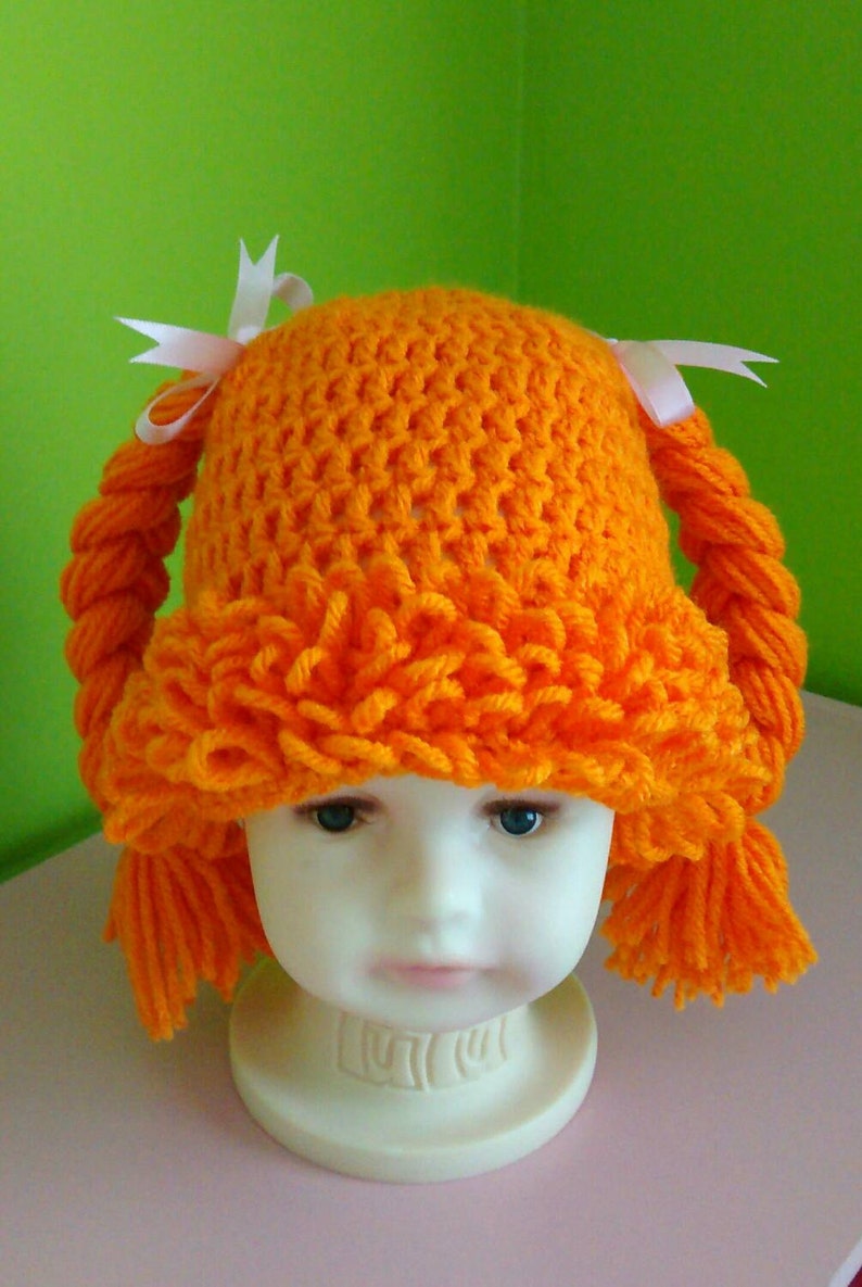 May include: Orange crocheted hat with two long braids and white bows. The hat is on a white mannequin head.