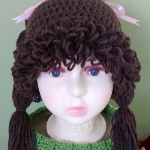 May include: A brown crocheted hat with pink ribbons and brown yarn hair. The hat is on a mannequin head wearing a green crocheted scarf.