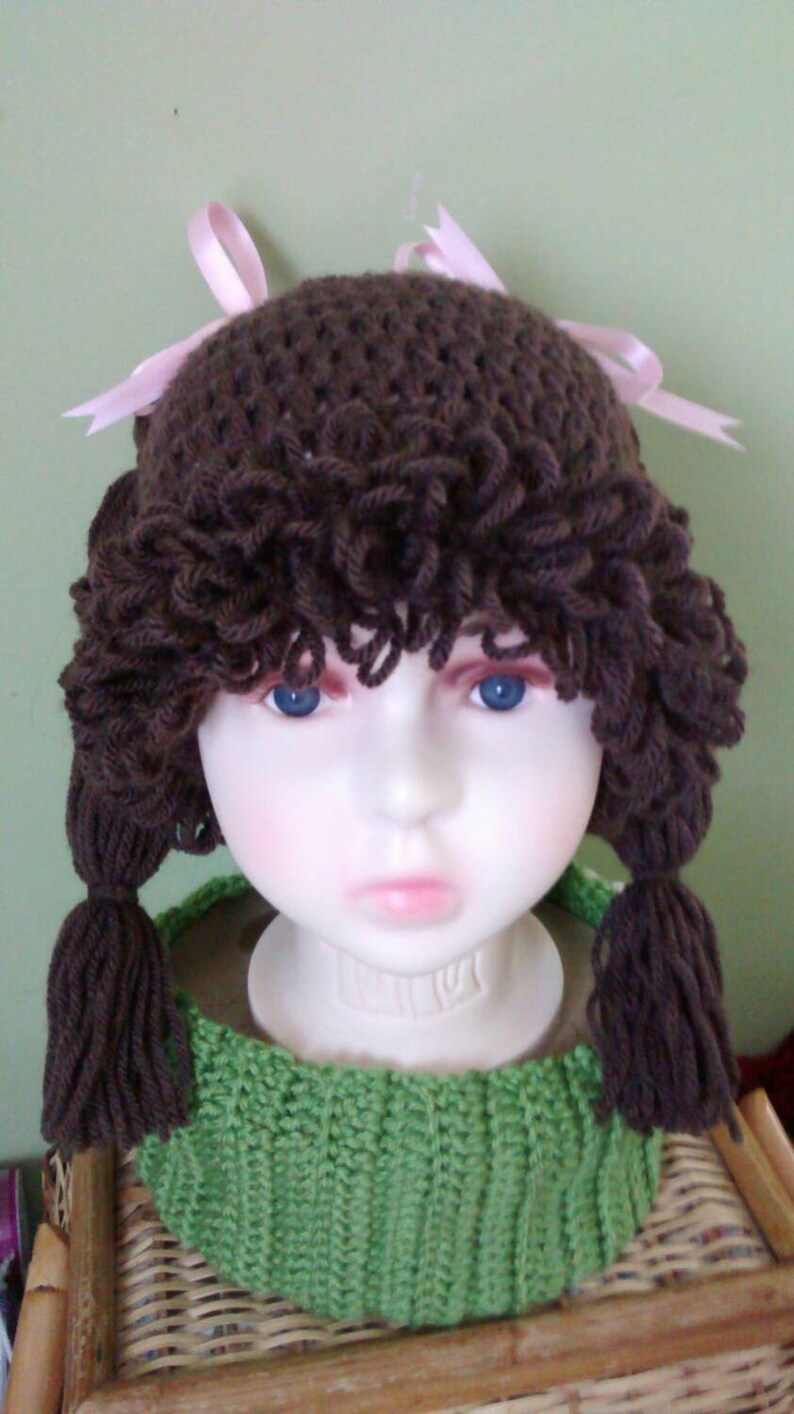 May include: A brown crocheted hat with pink ribbons and brown yarn hair. The hat is on a mannequin head wearing a green crocheted scarf.