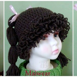 May include: A brown crocheted hat with long brown yarn pigtails and pink bows. The hat is on a mannequin head.
