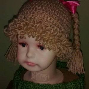 Pode incluir: Um chap&eacute;u de croch&ecirc; marrom com tran&ccedil;as e la&ccedil;os rosa. O chap&eacute;u est&aacute; em um manequim que est&aacute; usando uma echarpe de croch&ecirc; verde.