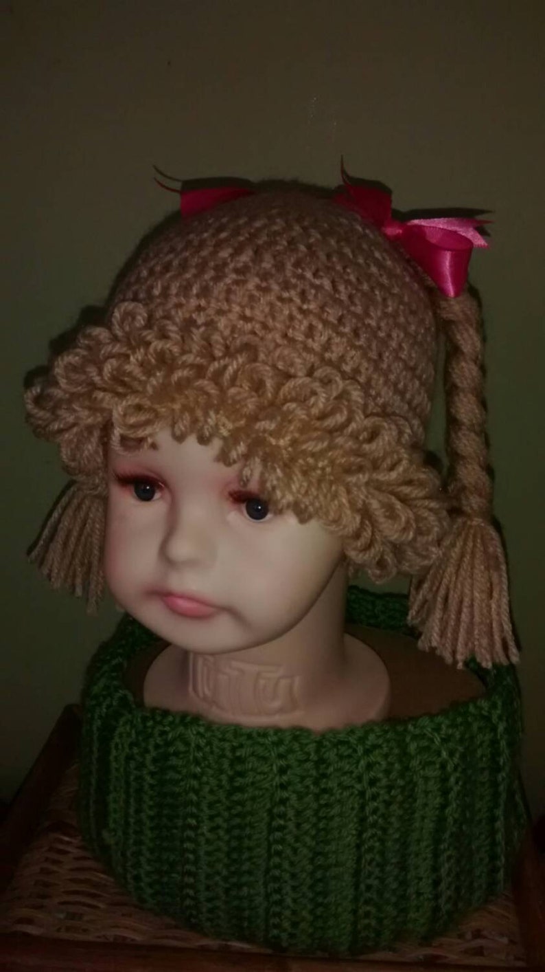 Pode incluir: Um chap&eacute;u de croch&ecirc; marrom com tran&ccedil;as e la&ccedil;os rosa. O chap&eacute;u est&aacute; em um manequim que est&aacute; usando uma echarpe de croch&ecirc; verde.