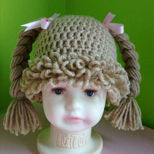 May include: A brown crocheted hat with two pigtails and pink bows. The hat is on a doll's head with blue eyes.