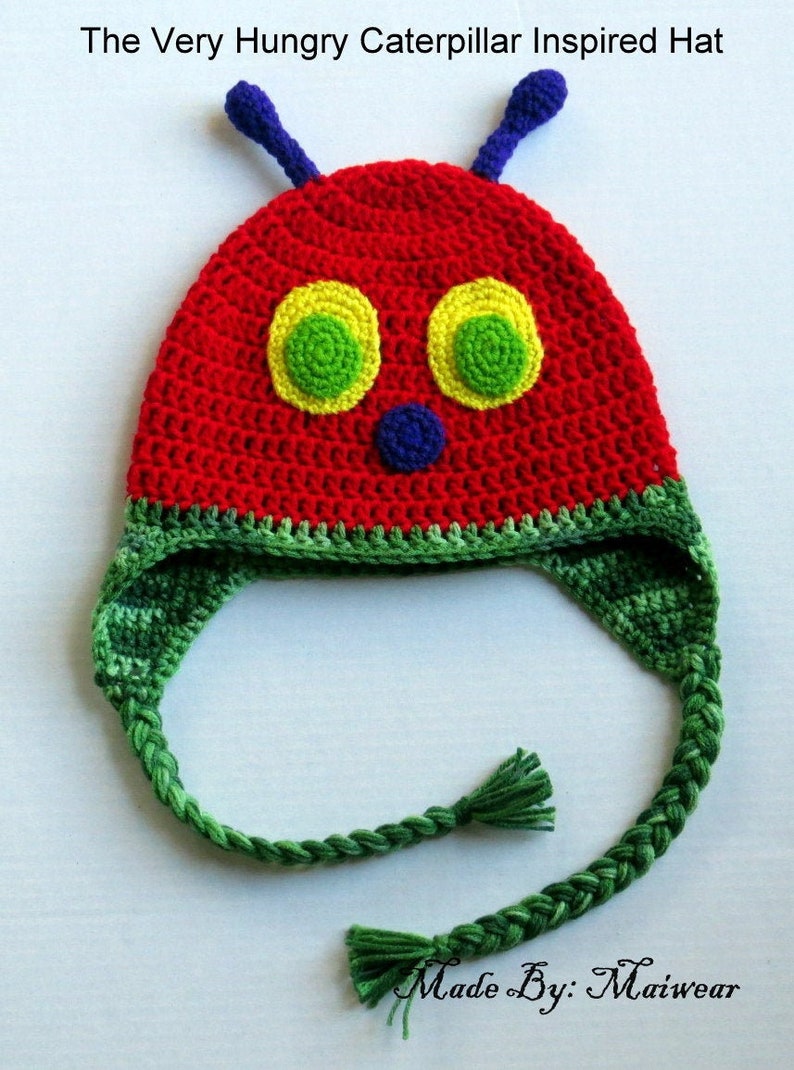 May include: A red, green, and yellow crocheted hat inspired by The Very Hungry Caterpillar book. The hat has a long green braided strap with tassels at each end. "Made By: Maiwear" is written on the white background.