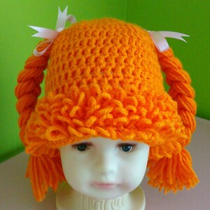 Pode incluir: Chap&eacute;u de croch&ecirc; laranja com duas tran&ccedil;as longas e la&ccedil;os brancos. O chap&eacute;u est&aacute; em uma cabe&ccedil;a de manequim branca.