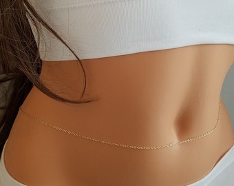 May include: A delicate gold-colored body chain. The chain is worn around the waist, creating a subtle, elegant accessory. The chain is thin and appears to be made of small, connected links. The model is wearing a white top and bottoms.