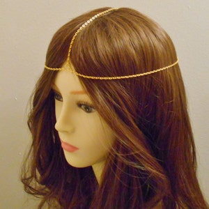 May include: A gold chain headpiece with a delicate design. The chain is worn on a mannequin with long, brown hair.