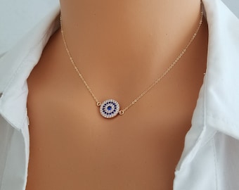 Könnte beinhalten: Goldkette mit einem blauen und weißen bösen Blick Anhänger. Der Anhänger ist von kleinen, klaren Steinen umgeben.