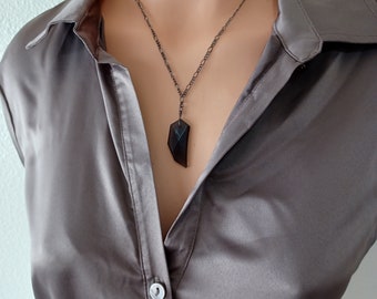 May include: A dark, geometric pendant necklace with a gunmetal chain. The pendant is a dark, faceted stone, suspended from a delicate chain. The necklace is displayed on a model wearing a gray satin blouse.