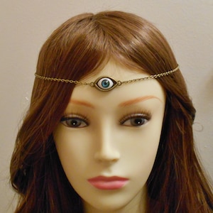 May include: A gold chain headband with a blue and white evil eye charm. The headband is worn on a mannequin with long, brown hair.