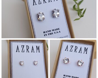 Puede incluir: Tres pares de pendientes en cajas pequeñas. Las cajas son marrones con texto blanco que dice "AZRAM" y "HAND MADE IN THE U.S.A". Los pendientes son plateados y dorados con piedras transparentes. El primer par es de forma ovalada, el segundo par es de forma de corazón y el tercer par es redondo.