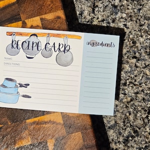 Op de afbeelding: Een witte receptenkaart met een blauwe en grijze illustratie van keukengerei. De kaart heeft de woorden "Recipe Card" in zwarte cursieve letters. De kaart heeft secties voor "Name", "Directions" en "Ingredients".