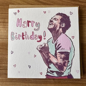 May include: A birthday card with the words "Harry Birthday!" in pink and green. The card shows an illustration of a person celebrating, with small hearts scattered around. The card is on a wooden surface.
