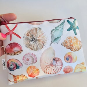 Puede incluir: Una bolsa blanca con cremallera con un estampado de acuarela colorido de conchas marinas y estrellas de mar. La bolsa tiene una cremallera granate y una pequeña etiqueta con la palabra "Gigi" en ella.