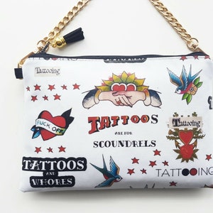 Op de afbeelding: Witte canvas clutch met een gouden kettingband. De clutch heeft verschillende kleurrijke tattoo-achtige ontwerpen, waaronder harten, zwaluwen en ankers. De tekst "Tattoos are for Scoundrels" staat op de clutch gedrukt.