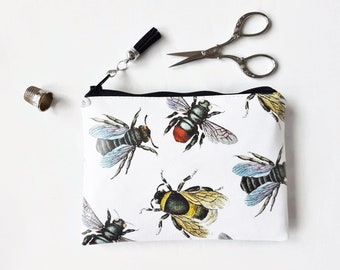 May include: White zippered pouch with a black zipper and a black tassel. The pouch has a repeating pattern of bees with black, yellow, red, and blue accents.