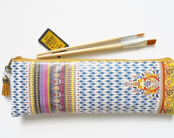 May include: A yellow and blue patterned zippered pouch with a tassel. Two wooden paintbrushes are laying on top of the pouch. A small yellow and black package labeled "Best Dorcas Needles 4/8 mm/prs" is also visible.