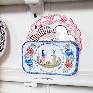 Op de afbeelding: Een blauw en wit rechthoekig dienblad met een geschilderd tafereel van twee mensen in traditionele kleding die op een veld met bloemen staan. Het dienblad heeft een blauwe en witte rand en staat op een witte plank.