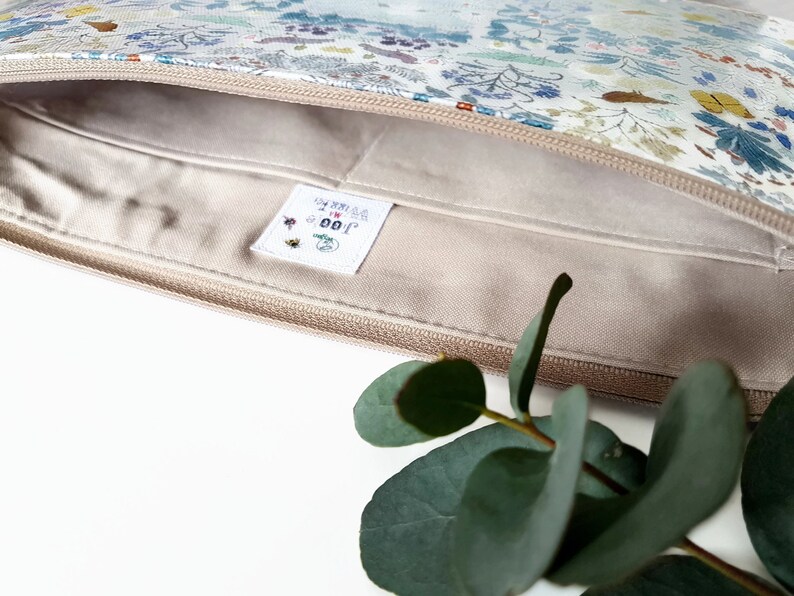 May include: A beige zippered pouch with a white floral print and a white label with the text "GOOD FOR YOU" in black and red lettering.