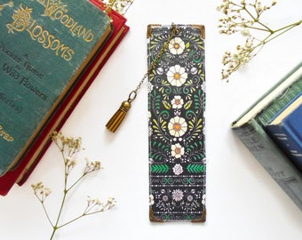 May include: A decorative bookmark with a floral pattern in white, yellow, and green on a black background. It has a gold tassel and corner protectors. The bookmark is next to vintage books.
