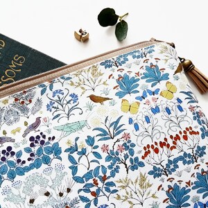 May include: A white zippered pouch with a floral and bird print. The pouch has a brown tassel zipper pull. The pouch is partially open and a green book with the text "Woodland Blossoms" is visible inside.