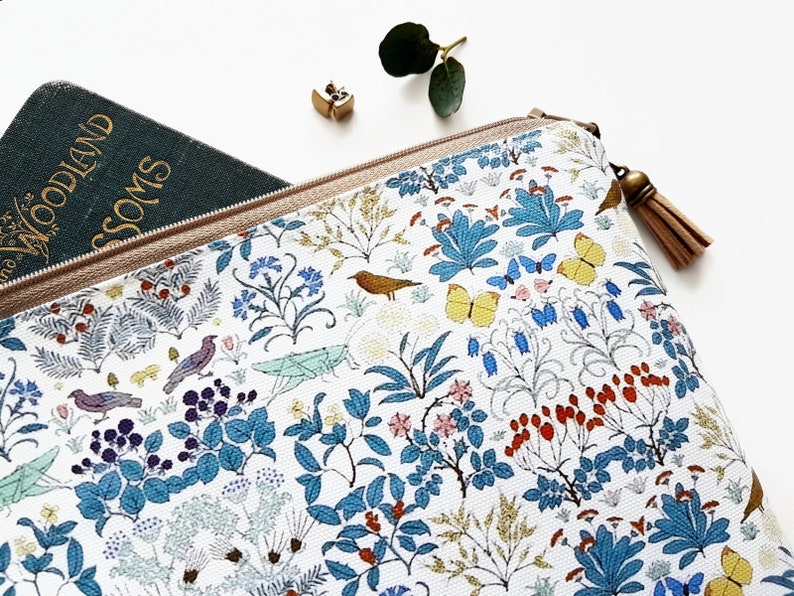 May include: A white zippered pouch with a floral and bird print. The pouch has a brown tassel zipper pull. The pouch is partially open and a green book with the text "Woodland Blossoms" is visible inside.